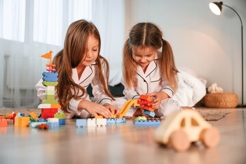 Naklejka premium Bunch of plastic construction toys are on the floor. Two little girls are playing and having fun together in domestic room