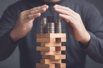 Risks in business or financial concept. Idea to prevent risk in business. Business man using hand to protection coin stacked on tower wooden block game and prevent falling down.