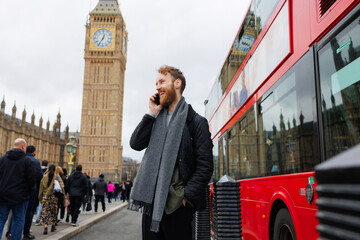 30s bearded man talking on the phone near big ben next to a passing red bus