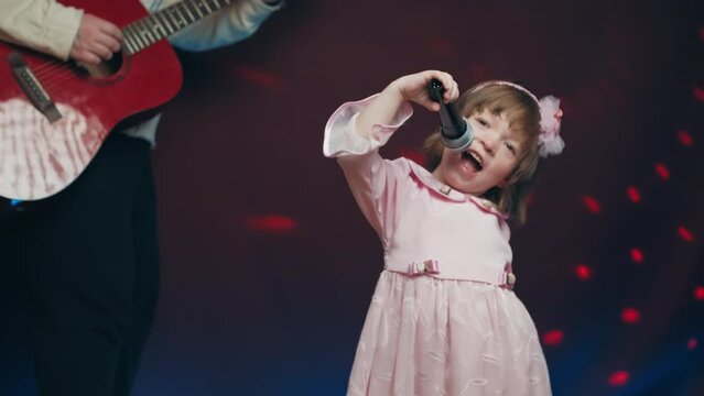 Little girl on stage in vintage dress, she sings into microphone and dances, her father plays acoustic guitar. Color music is shining. Performance on stage. Early development of children