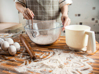 woman in an apron in the kitchen prepares dough from flour, flour, eggs and a glass transparent bowl , close up, cooking at home
