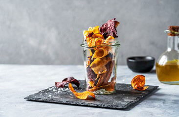 Healthy vegetable chips in a glass jar on stone cutting board with marine salt and oil aside....
