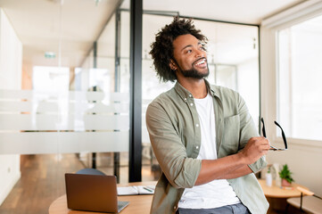 Successful cheerful african-american male entrepreneur, small business owner, male office employee, black businessman wearing green casual shirt stands in confident pose with arms crossed and laughing