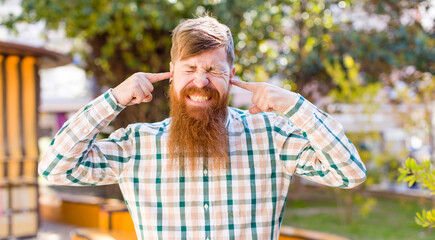 red hair bearded man looking angry, stressed and annoyed, covering both ears to a deafening noise, sound or loud music