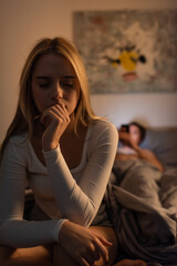worried woman sitting on bed near blurred boyfriend in bedroom, cheating concept.