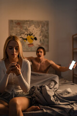 Obraz premium blonde woman sitting on bed next to screaming boyfriend holding smartphone in bedroom, cheating concept.
