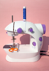 Mini electric sewing machine with a spool of blue thread and small scissors. Tailoring equipment isolated on pink. Cute stylish sewing machine toy for children and beginners