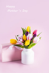 a bouquet of colorful tulips in a modern white vase on a pink background with a gift box. the text of a happy mother's day. vertical view.