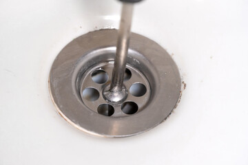 Obraz premium Close-up of a screwdriver unscrewing a drain in a white sink. Jack of all trades, repairing or cleaning a clogged sink drain in the bathroom. Concept of maintenance of plumbers.