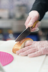 The cook cutting bun for hot dog in Greek fast food restaurant. Chef using sharp Japanese santoku knife to cut bread