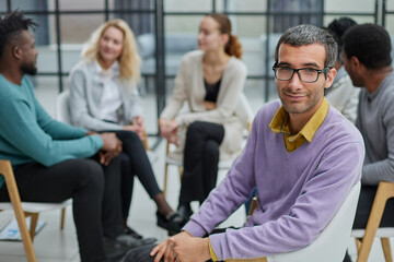 A happy young businessman, in a purple sweater, sits against the background of his colleagues
