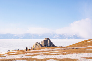 Frozen Baikal Lake. Olkhon Island in winter. View on famous Shamanka Rock, an old larch tree on hill and tourists traveling on ice in cars. Winter holidays