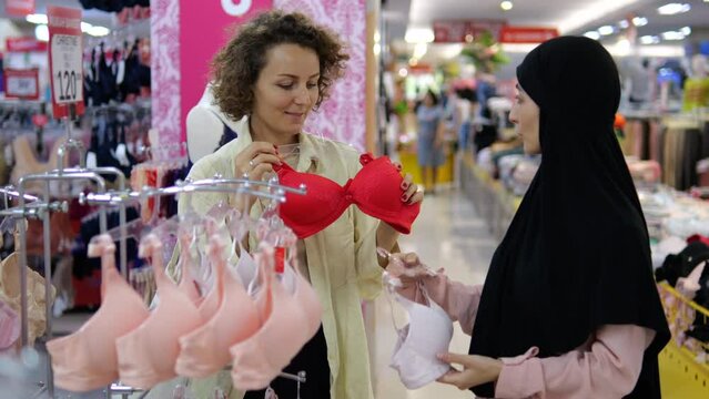 Two friends consult, choose and try on bras in mass-market store in shopping center. Caucasian and Armenian woman communicate and discuss, choosing purchase. Two girls girlfriends in mall on shopping.