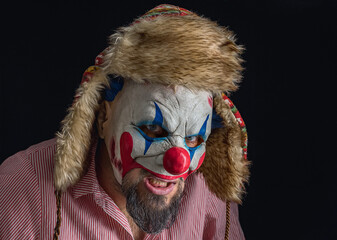 A bearded man wearing a creepy clown mask and a hat with ears looks into the camera. Black...