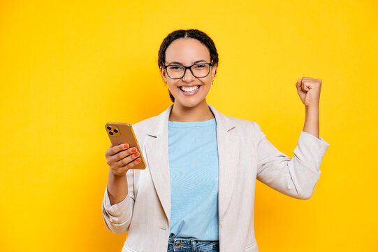 Joyful happy mixed race woman with glasses, holding her smartphone in hand, rejoices in success or win, looks at camera, make YES gesture, stand on isolated yellow background