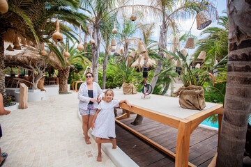 A mother and daughter enjoying their summer vacation together, smiling in front of a beautiful garden by the swimming pool.