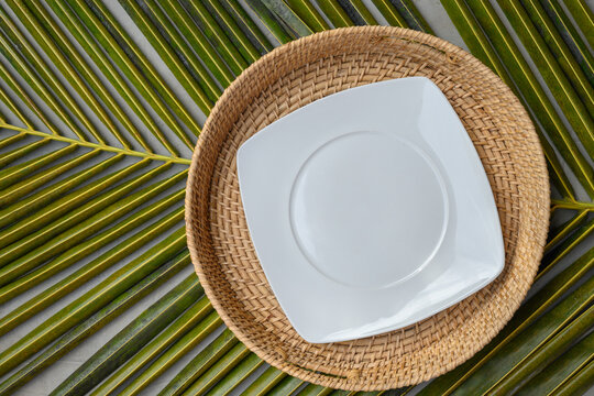 Empty square white plate in authentic wicker tray on green palm leaf. View from above. Space for design or text. Template for Asian and tropical cuisine.