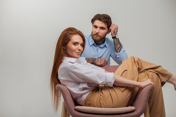 Fashion couple of lovers. Beautiful happy redhead girl model with red hair sits and rest in a chair and handsome brutal hipster man with beard and tattoo