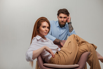 Beautiful fashionable young couple in trendy clothes in the studio. Pretty red-haired girl in a white shirt sits in a chair next to a handsome hipster man with a tattoo and a beard