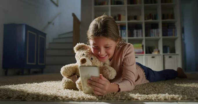 Portrait of a Little Female Child Hugging her Teddy Bear and Watching a Video on a Smartphone in the Living Room. Happy Cute Girl Using Phone, Playing Mobile Educational Games. Concept of Childhood