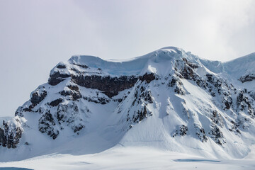 Rugged mountain peak on Antarctic Peninsula. Steep slopes covered with snow; underlying rocks exposed. Blue ice visible. Cloudy sky in background.
