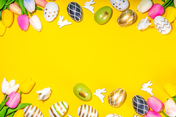 Bright Easter greeting card background. Trendy colored black, golden, stripe painted Easter eggs with white bunny rabbits decor, tulip flower on high-colored yellow background, flat lay copy space