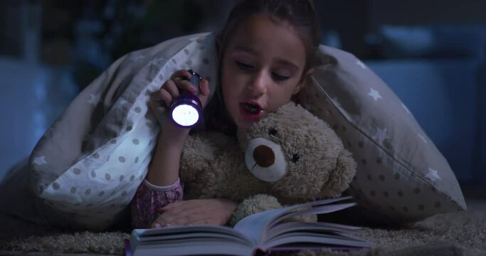 Portrait of Little Female Child Reading a Book to her Favorite Teddy Bear Before Sleeping. Happy Cute Girl Playing with her Toy, Using Flashlight, Telling a Story. Concept of Childhood and Imagination
