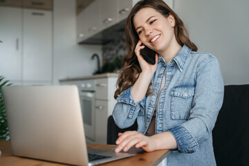 Young woman sits at the kitchen table using a laptop and talking on a cell phone