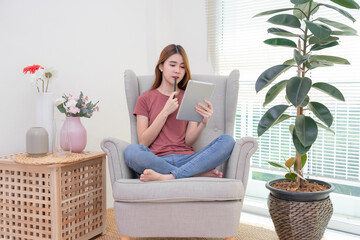 Asian woman sitting on yellow sofa using digital tablet and pen for learning shopping or working online near window