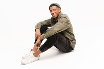 Full length photo of carefree smiling african-american guy in hat sitting on the floor in relaxed...