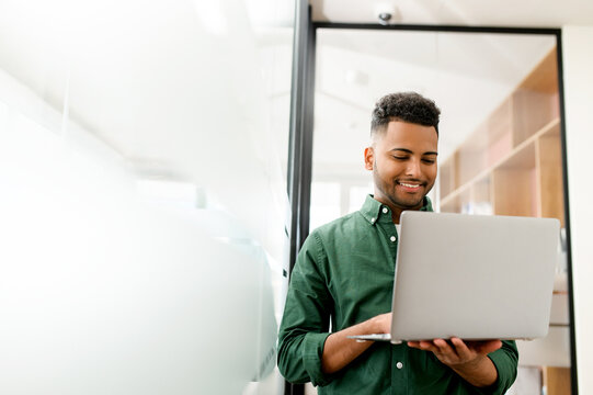 Portrait of charismatic elegant young indian businessman standing in the modern office and using laptop, arab male freelancer is enjoying work from coworking space