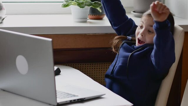 An emotional portrait of a little girl sitting at a computer and joyfully raising her hands up with a shout of joy of victory.