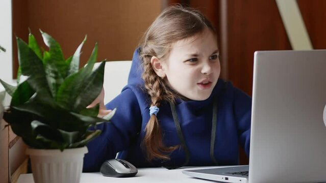 Portrait of a small, stupid Caucasian girl with pigtails sitting in front of a laptop and indignantly saying something, wrinkled her face.