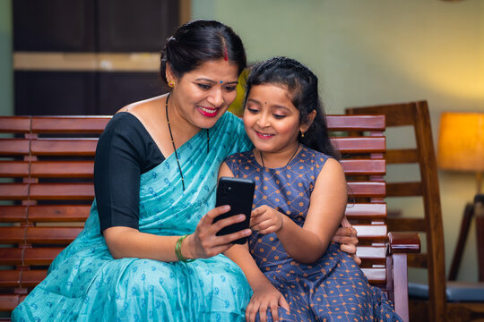 Happy mother showing mobile phone to daughter while sitting on sofa at home - concept of technology, bonding and motherhood.