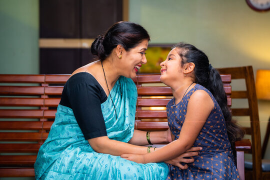 Happy mother playing with daughter by cuddling while sitting on sofa at home - concept of motherhood, family bonding and weekend holidays.