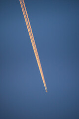 Passenger jet plane flying at high altitude leaving golden trail of smoke contrails from the sunrise. Close up telephoto view taken from ground level. - 580750407