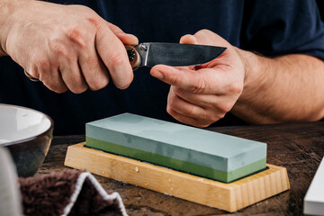A man sharpens a knife with a grindstone on a rustic wooden table.
