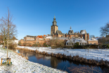 Canal and medieval wall of Hanseatic Dutch tower town Zutphen in The Netherlands covered in snow with historic heritage buildings against a clear blue sky. Winter wonderland picturesque scene.