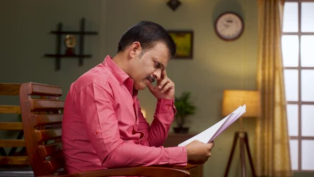 Thoughtful worried man holding paper documents while sitting on chair at home - concept of financial issues, debt notice and