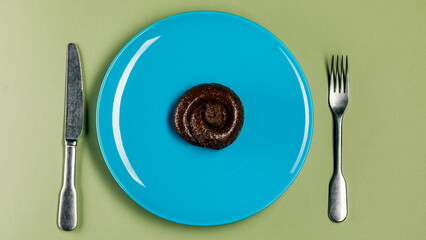 Plastic excrement lies on a plate between a fork and a knife close-up. The concept of an inept cook, fake news, shitty restaurant, bad food, taste of shit