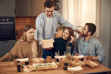 Man is standing. Friends are sitting indoors at home and eating sushi together
