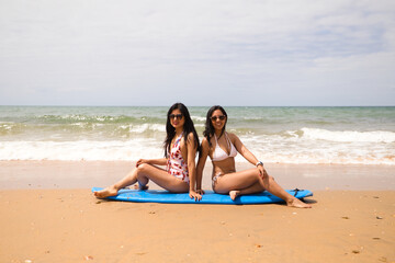 Two young and beautiful women sitting on a surfboard on the shore of the beach. The women are enjoying their trip to the beach paradise. Holidays and travels.
