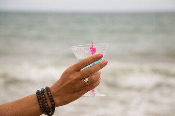 Detail of hand raising glass in the air with a blue drink. In the background you can see the sky and the sea in the horizon.