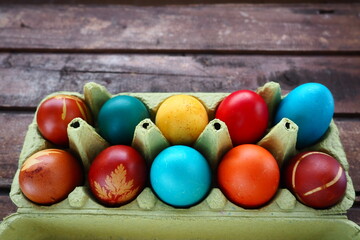 Multi-colored eggs painted with gouache and onion peel in a cardboard box on a wooden table background. Ten boiled eggs. Postcard poster for Easter. Easter holiday. Red, blue, yellow, brown eggs