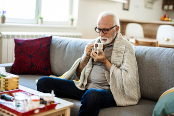 Senior man having a flu, resting in the bed and drinking tea. Sick senior man with fever wrapped in...