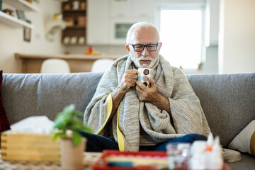 Senior man having a flu, resting in the bed and drinking tea. Sick senior man with fever wrapped in...