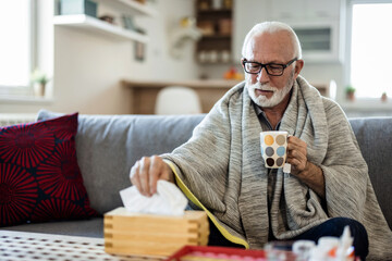 Covid, tea and sick senior man with a blanket and medicine on his living room sofa at home. Medical, health and wellness elderly person taking care of after a cold or flu sickness.