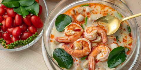 Tom Yam Kung or Tom Yum soup is Thai hot spicy soup shrimp or prawn with lemon grass, lemon, galangal, mushrooms, coconut milk and chili in bowl on wooden table top view, famous Thai food cuisine.
