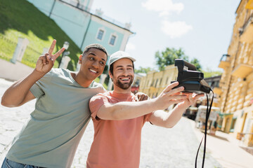 smiling african american man showing peace sign near bearded friend taking selfie on vintage camera on Andrews descent in Kyiv.