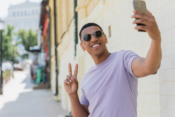 Smiling african american man in sunglasses showing peace sign while taking selfie on blurred urban...
